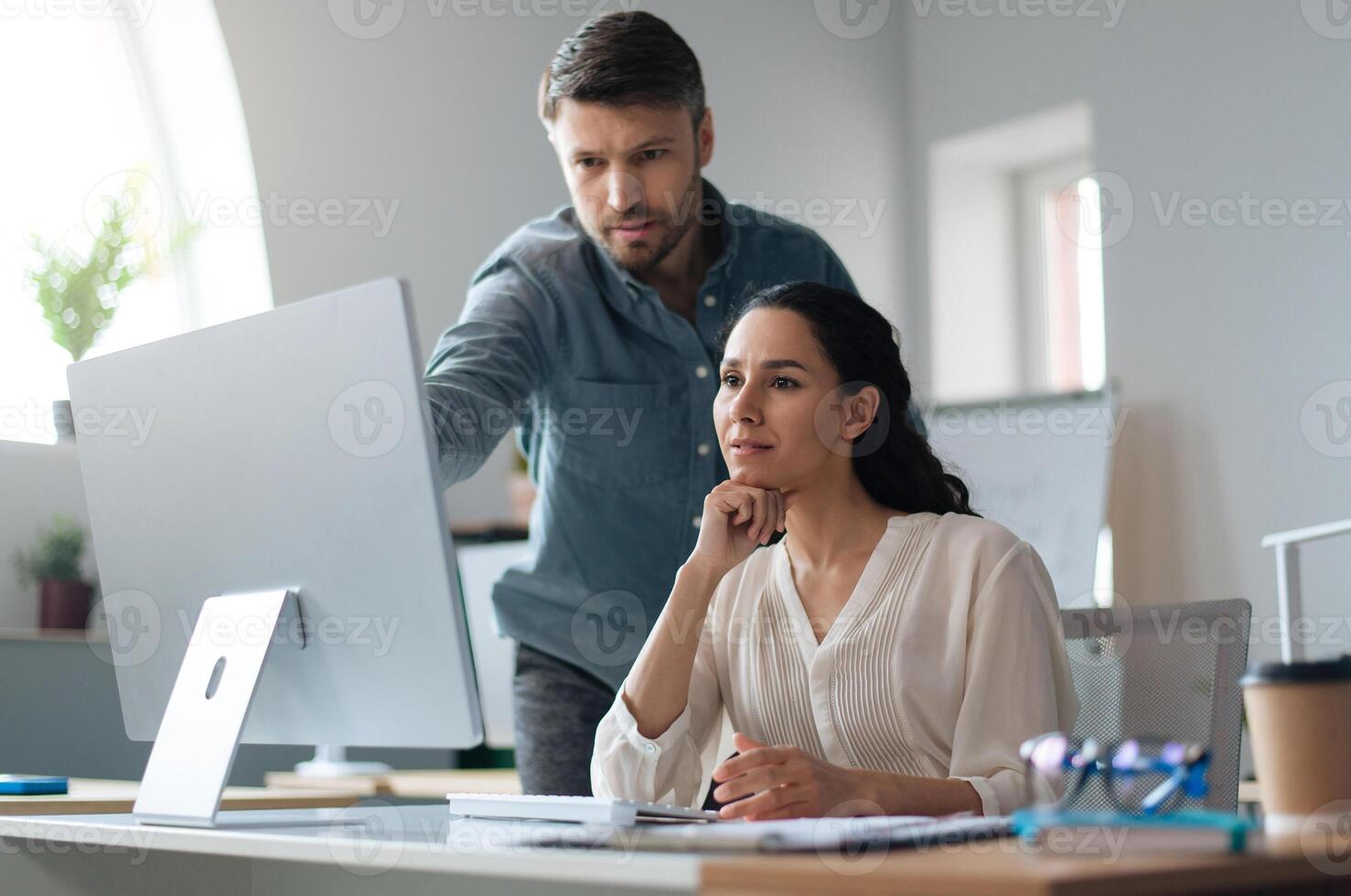Young female trainee working on computer together with her business mentor at modern company office. New team member on probation communicating to experienced male colleague photo