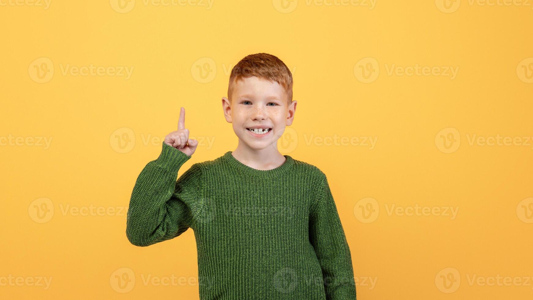 Happy ginger kid cute school boy in knitted green sweater pointing finger up over yellow studio background. Smiling redhead boy aiming up, having great idea or found creative solution photo