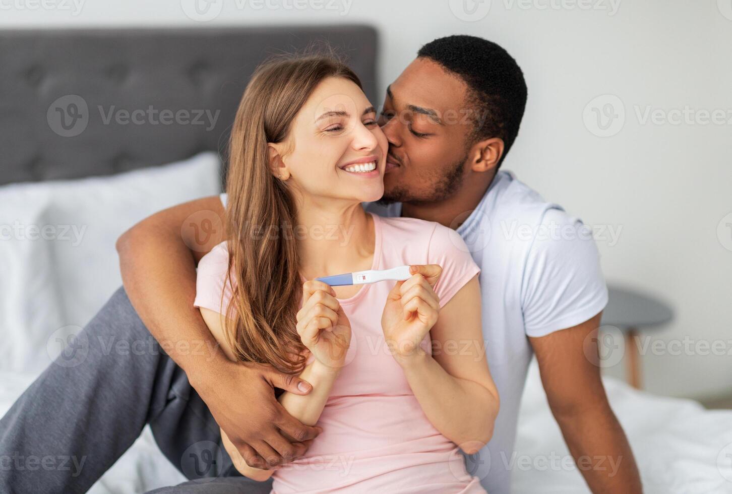 Happy multinational couple celebrating positive result of pregnancy test on bed at home. Young black man hugging and kissing his wife, overjoyed with future parenthood photo