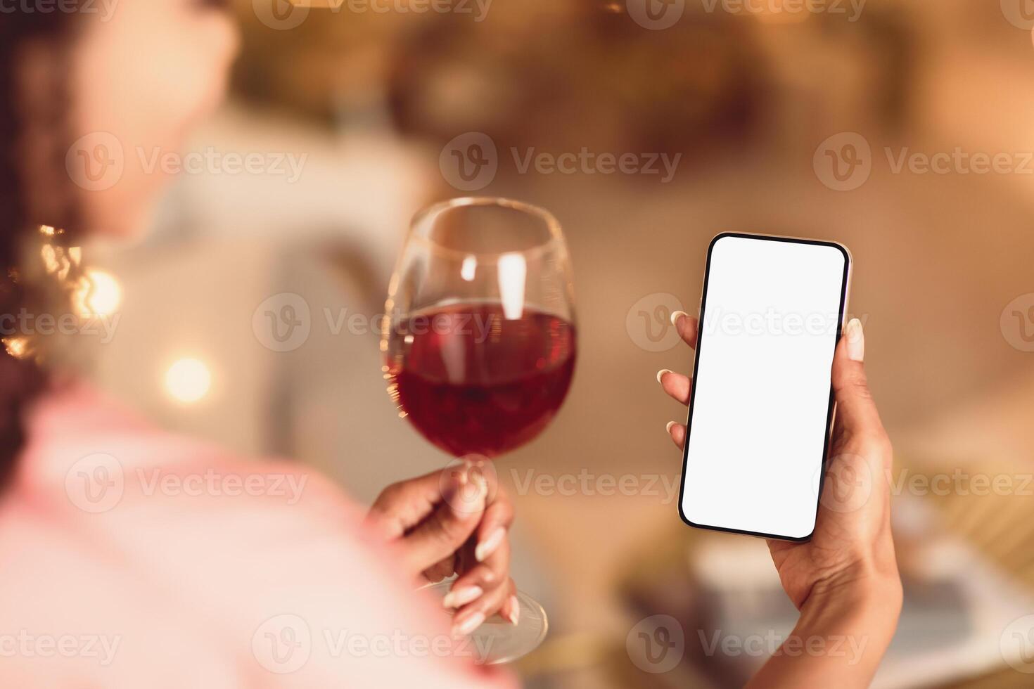 Back Over The Shoulder Back View Of Young Black Woman Holding And Using Mobile Phone With White Empty Screen For Mock Up, Making Virtual Call Or Watching , Drinking Red Wine From Glass photo