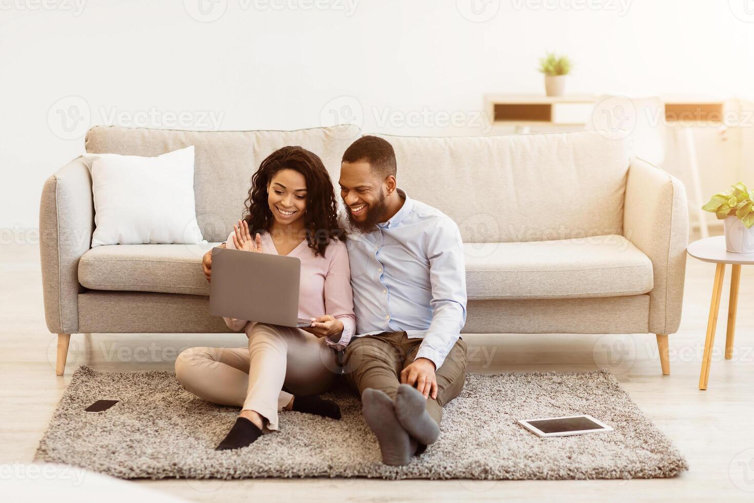 Virtual Communication. Happy black couple having online conference with friends or family using computer, waving hand to web camera, sitting on the floor carpet in bedroom at home photo