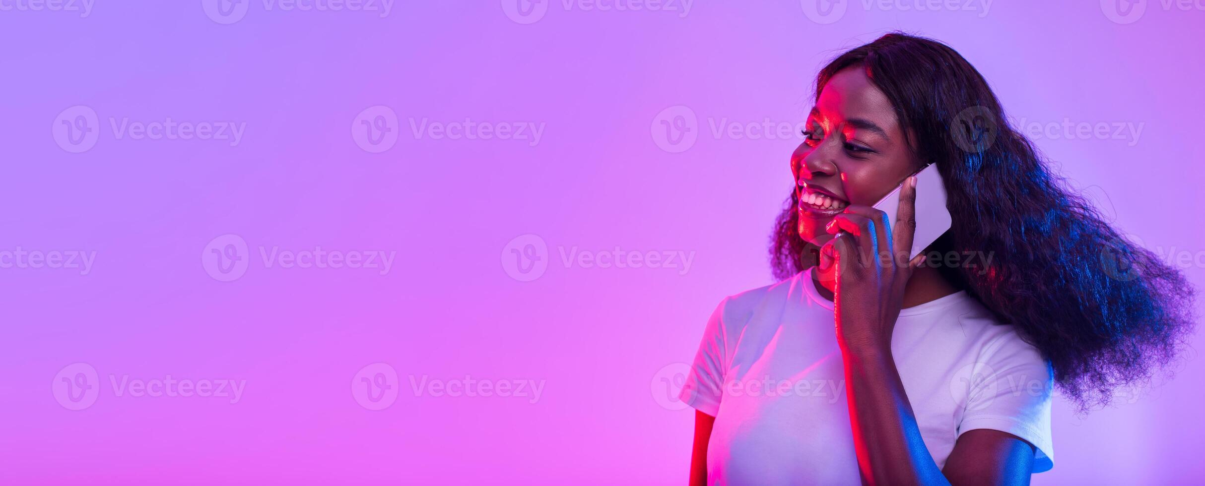 Smiling young African American woman having conversation on cellphone in neon light, banner design with copy space. Beautiful black lady talking with client or friend on smartphone photo