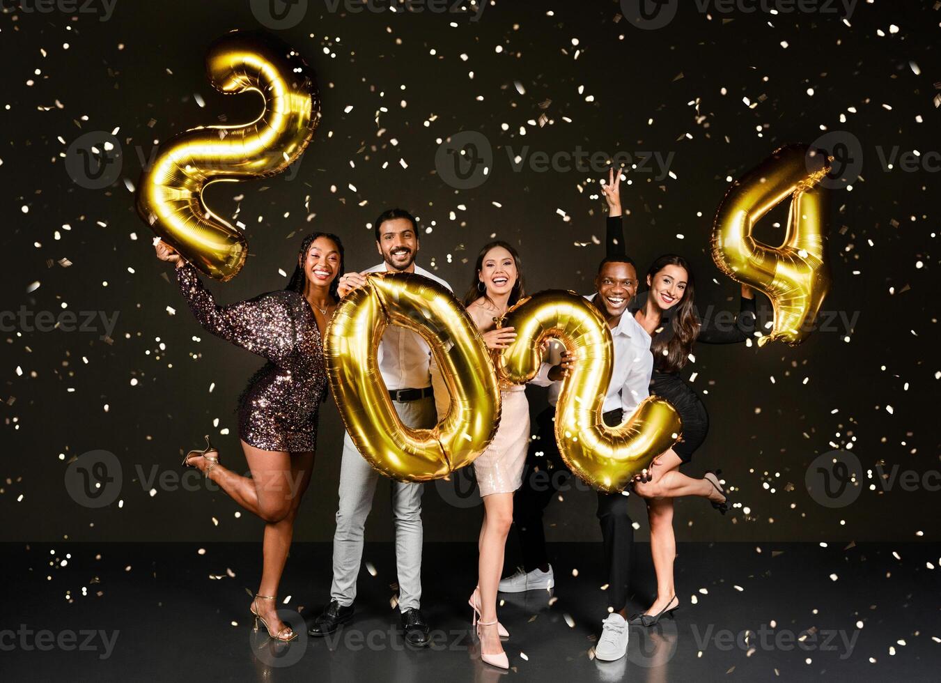 Happy beautiful multiethnic millennials men and woman in nice outfits celebrating New Year 2024 together, holding golden number balloons and posing over black background among confetti photo