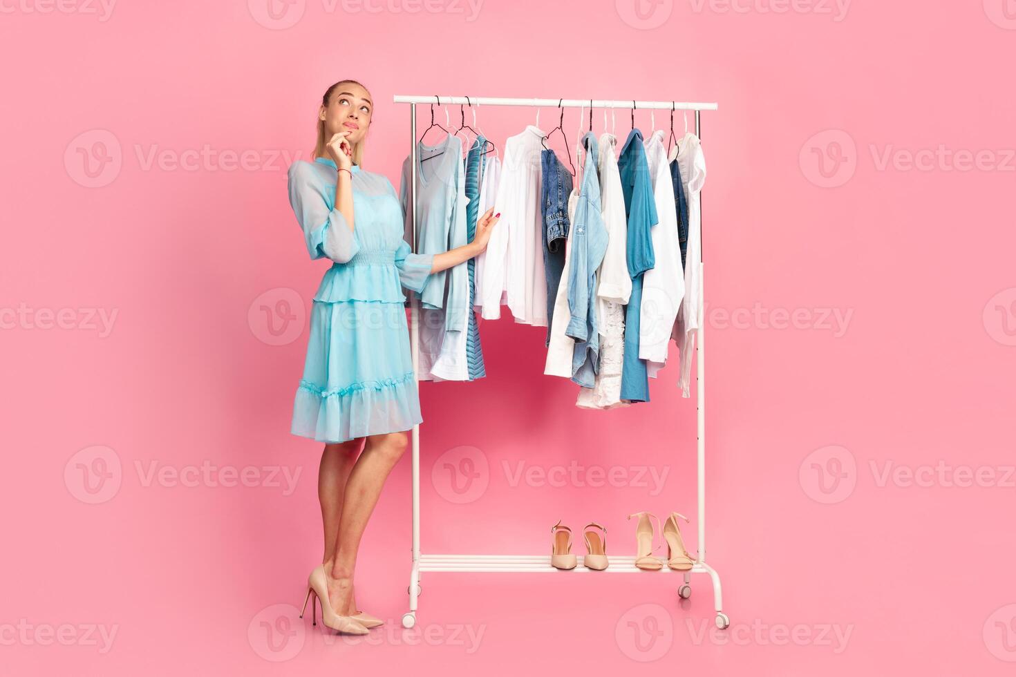 Making Choice. Vertical full length portrait of young pensive lady choosing trendy outfit, thinking about purchase and what to wear, standing in wardrobe near clothing rail, pink background photo