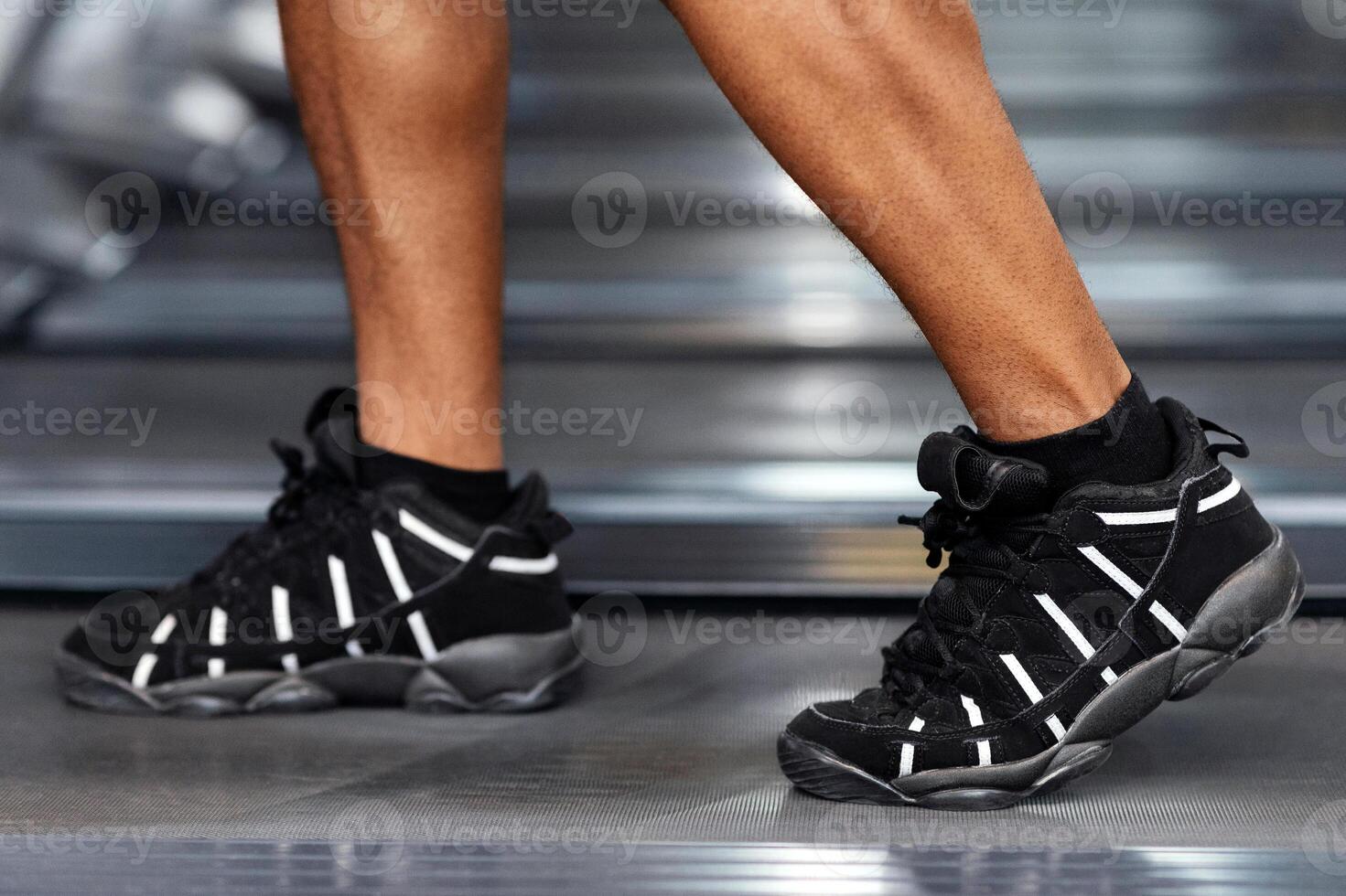 Sports shoes for gym concept. Closeup of unrecognizable african american man bodybuilder legs walking by treadmill, wearing black and white sneakers, selective focus on shoes, gym interior, copy space photo
