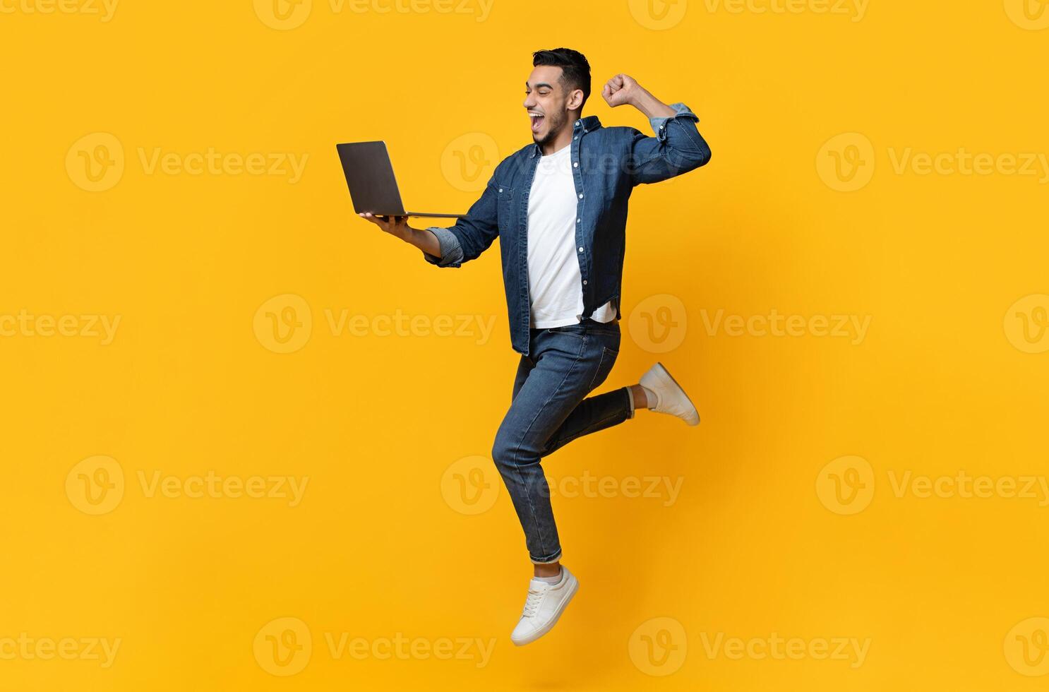 Emotional arab young man jumping up, using new modern laptop, raising fist up and smiling over yellow studio background, trading online, copy space for text or advertisement, panorama photo
