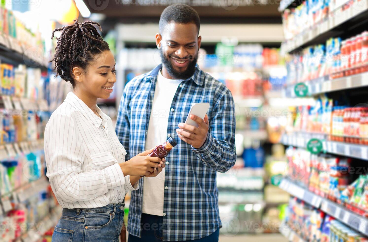 Black Family Doing Grocery Shopping In Supermarket. African American Couple Using Phone Scanning Food Product Standing In Store Aisles Indoors. Customers Buying Supplies In Local Shop photo