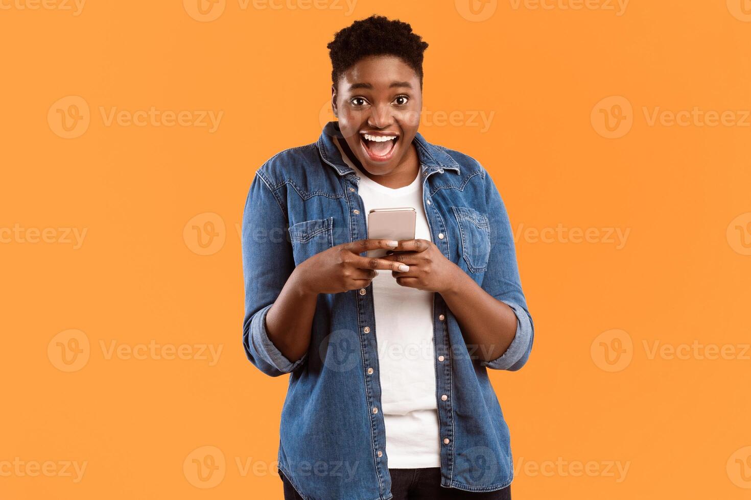 Great Application. Joyful Black Female Using New Smartphone And Mobile App Posing Smiling To Camera Standing Over Yellow Studio Background. Woman Playing Games On Phone Concept photo