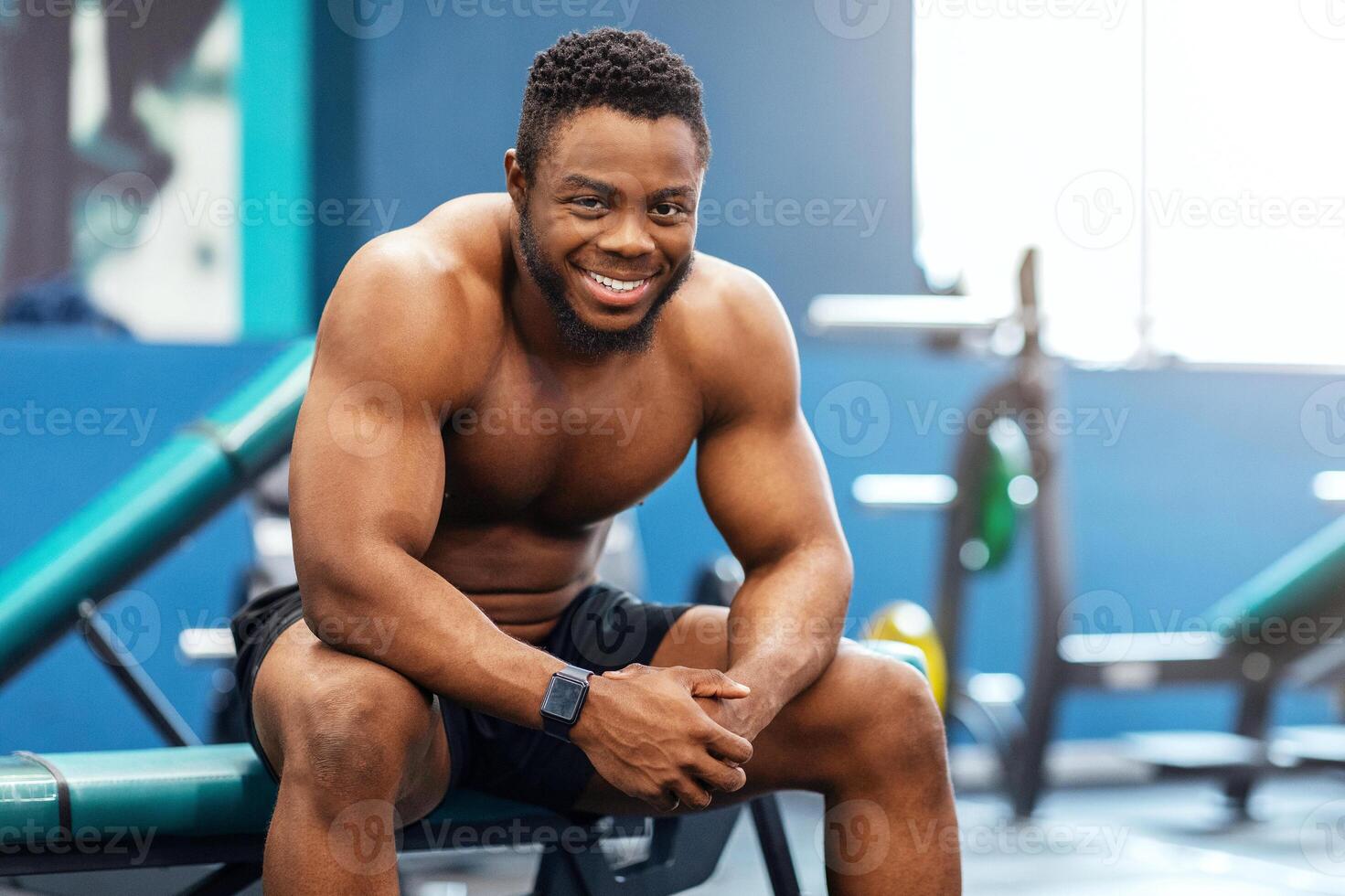 Smiling african american shirtless bodybuilder having break after workout at modern gym, smiling at camera. Happy black guy sportsman resting after exercising, sitting on bench, copy space photo