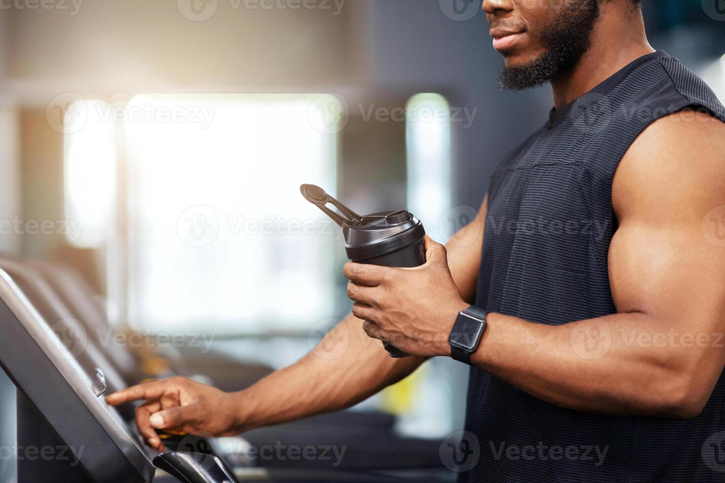 Unrecognizable black guy sportsman with protein drink standing on treadmill, side view, copy space. Cropped of muscular african american man having jogging work out, drinking water, sun flare photo