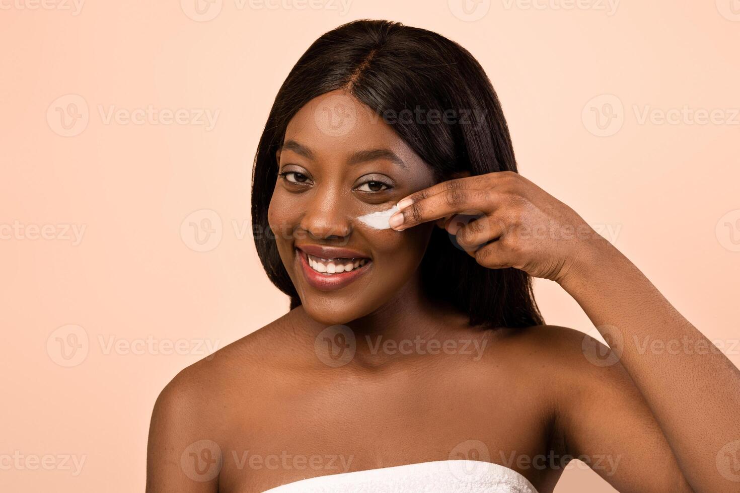 Delighted african american young pretty naked lady wrapped in towel with eye cream posing on beige studio background, closeup, eye care morning or evening routine concept, copy space photo