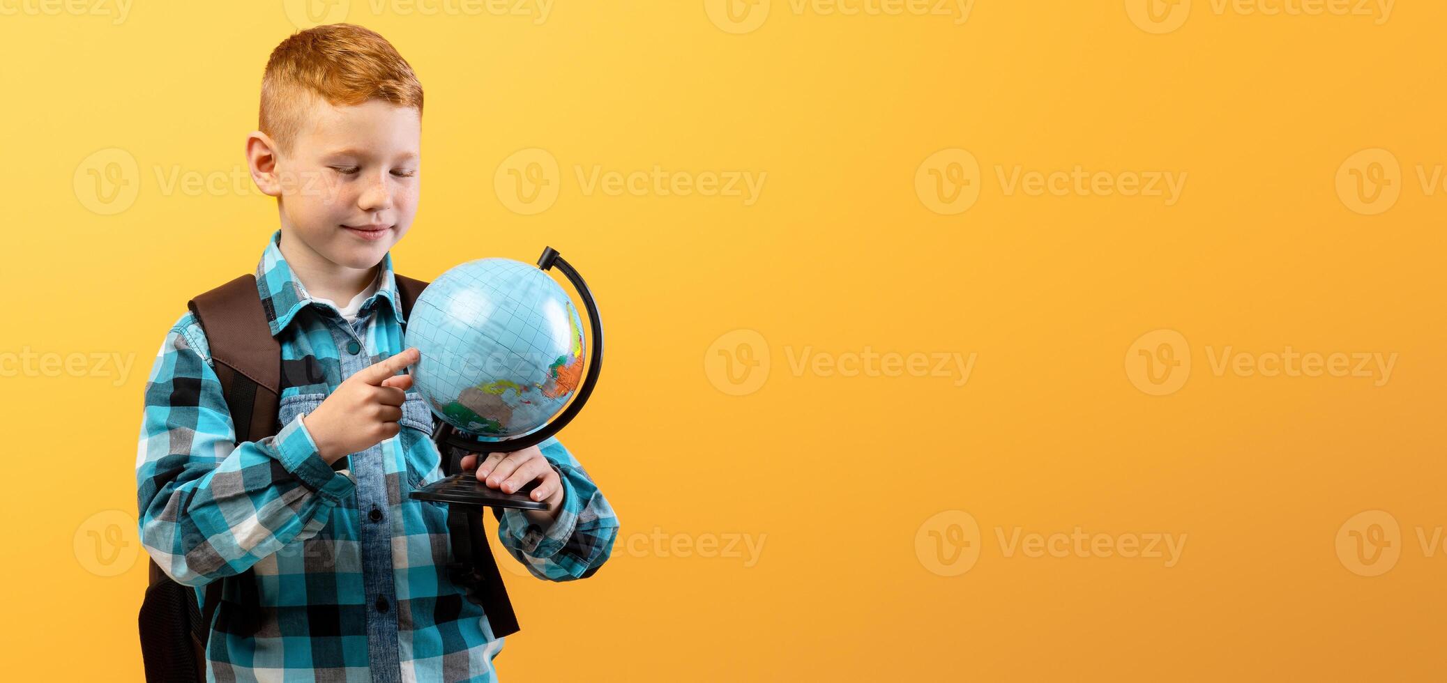 Positive schoolboy with backpack holding globe, studying geography on yellow studio background, copy space. Happy boy going to school, enjoying his school days. Kids education concept photo