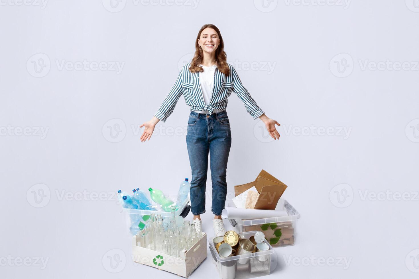 Waste Sorting. Happy woman standing among different kind of sorted garbage, showing boxes and containers with plastic, metal cans, glass bottles and paper for recycling over light studio background photo