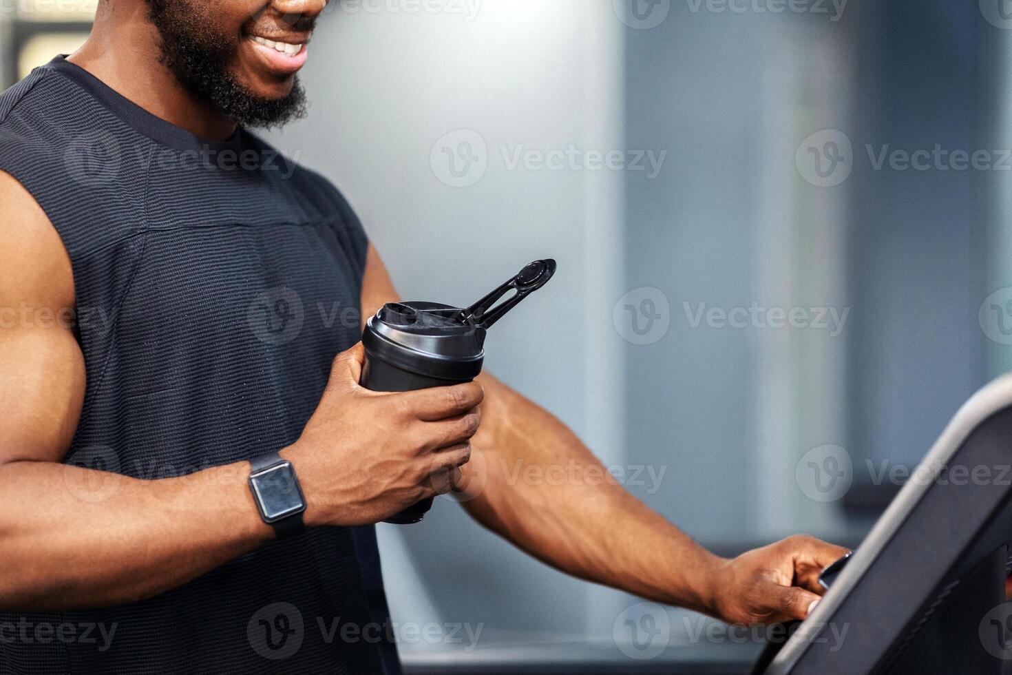 Cropped of muscular african american man having jogging work out, pressing on button. Unrecognizable black guy sportsman with protein drink standing on treadmill, side view, copy space, closeup photo