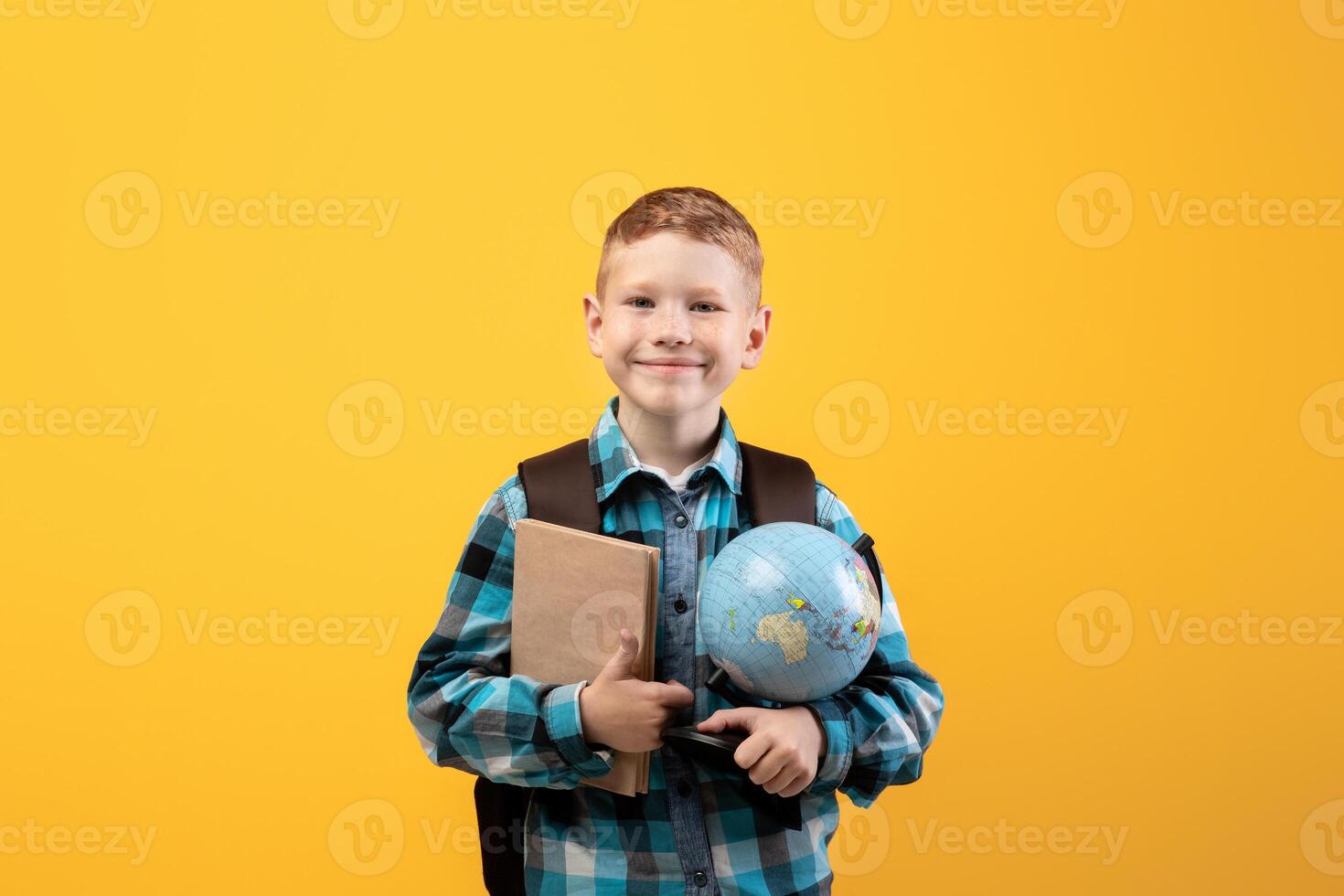 Positive schoolboy with backpack holding heap of books and globe, yellow studio background, copy space. Happy boy going to school, holding books, enjoying his school days. Kids education concept photo