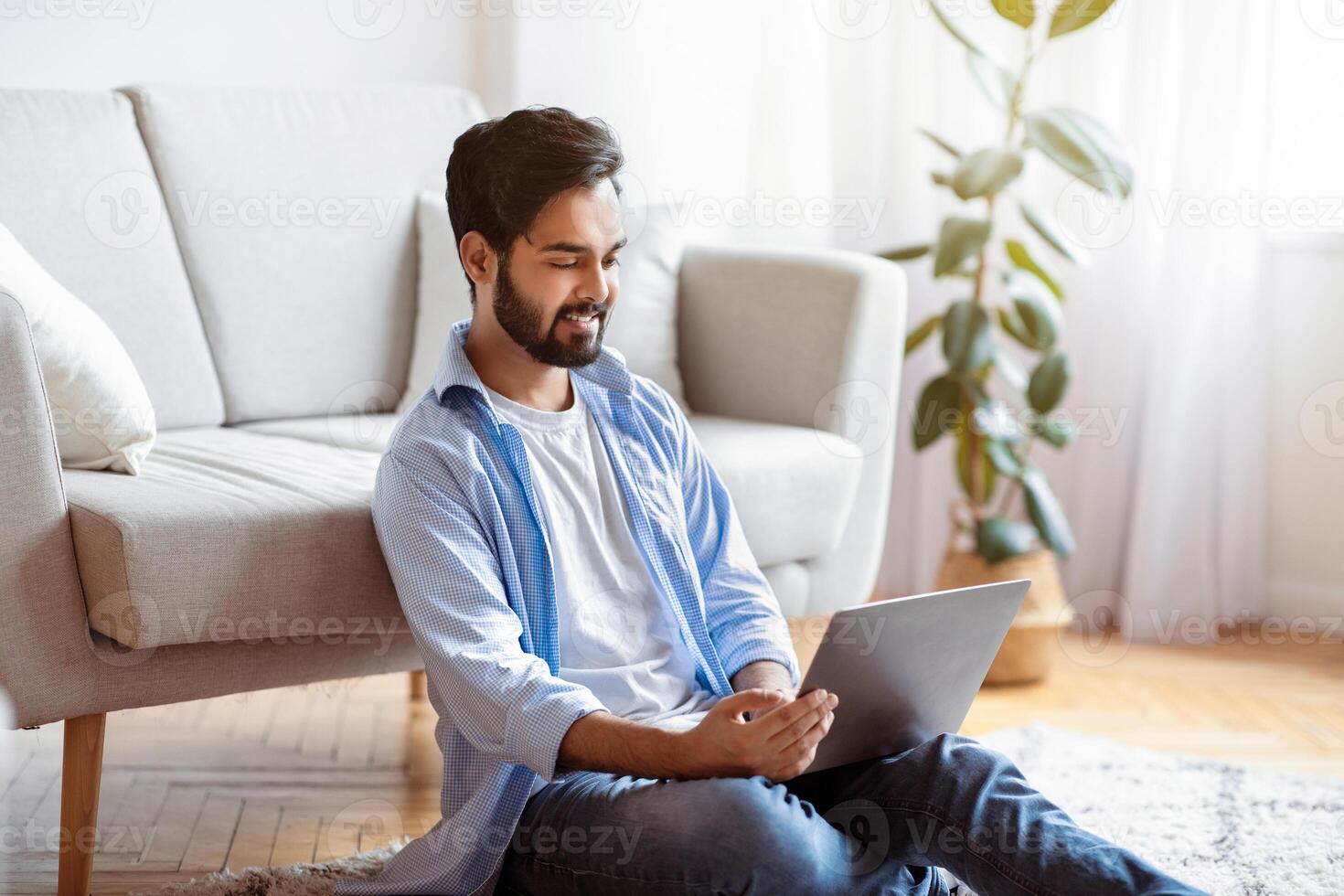 Telecommuting Concept. Young Eastern Guy Freelancer Working On Laptop While Sitting On Floor In Cozy Living Room At Home, Smiling Millennial Arab Man Enjoying Remote Job, Free Space photo