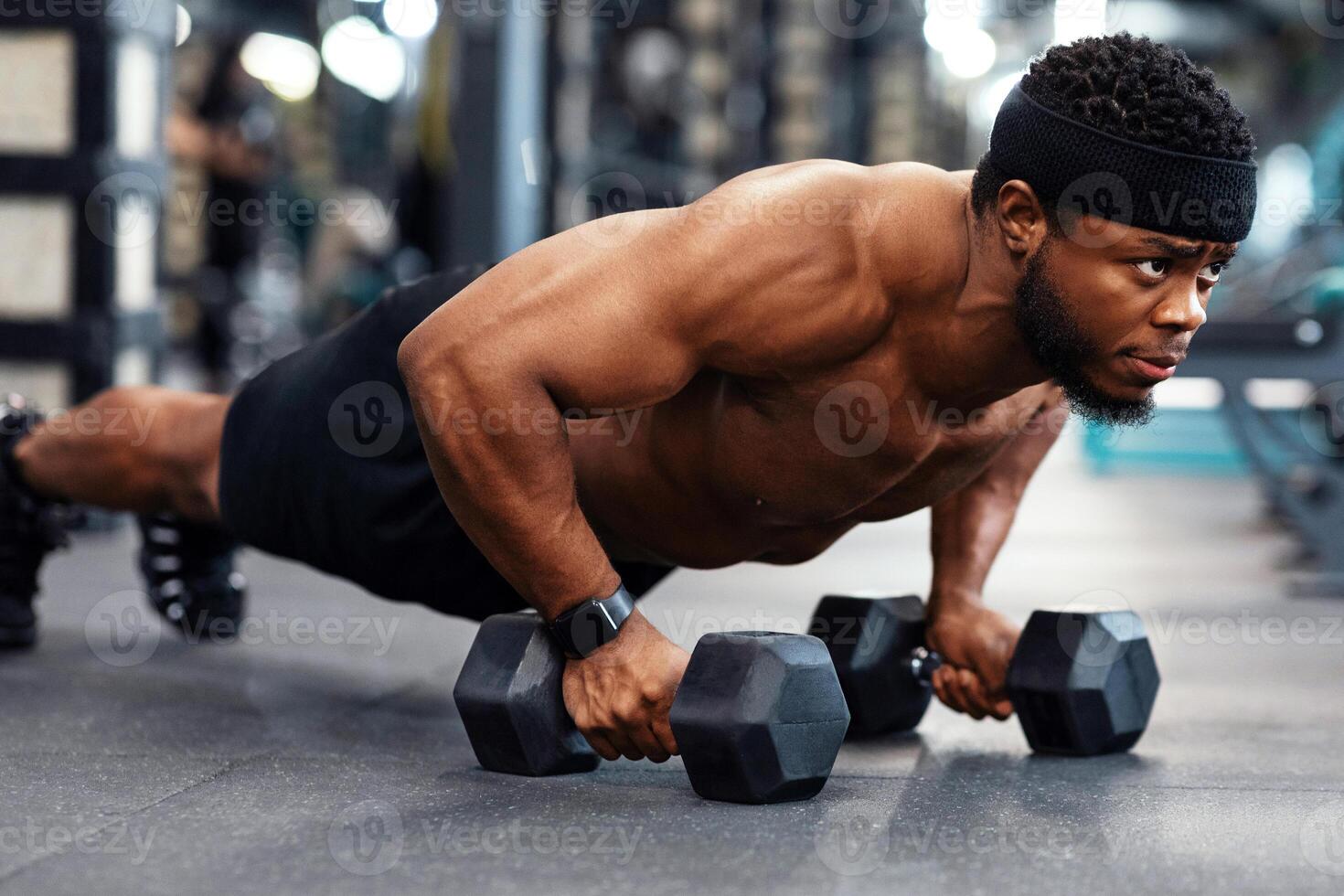 Concentrated black guy sportsman pushing up with dumbbells. Muscular african american bodybuilder shirtless man holding barbells and making push-ups from floor, work out at gym photo