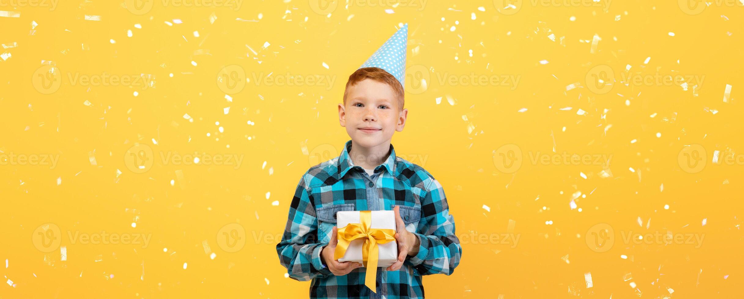 Smiling birthday boy in paper cap holding present, yellow studio background, copy space. Cute redhead kid celebrating his birthday, holding gift box, wondering whats inside photo