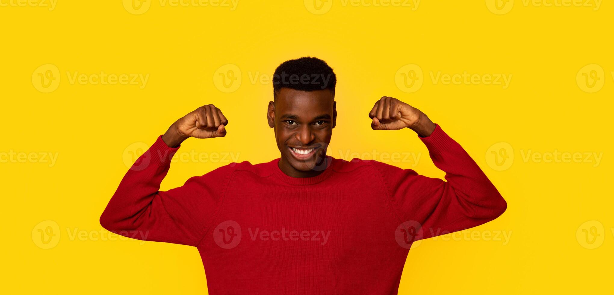 Funny Strong African American Guy In Red Sweater Showing Biceps Muscles And Looking At Camera, Cheerful Young Man Demonstrating His Strength, Standing Isolated Over Yellow Background, Copy Space photo