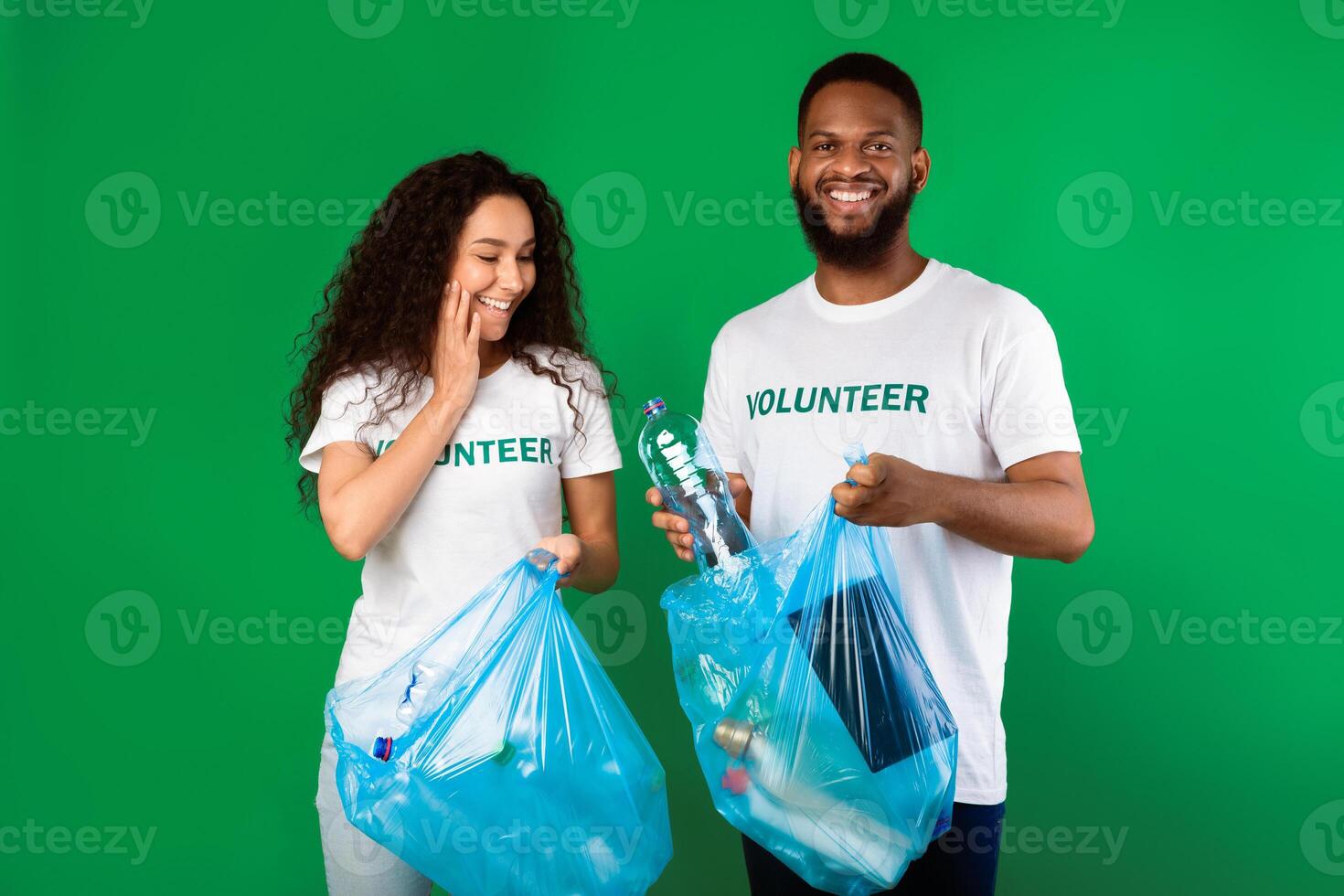 Couple Of Multicultural Volunteers Picking Plastic Litter And Bottles In Bags Cleaning Environment Standing Over Green Studio Background, Smiling To Camera. Volunteering Work And Ecology Concept photo
