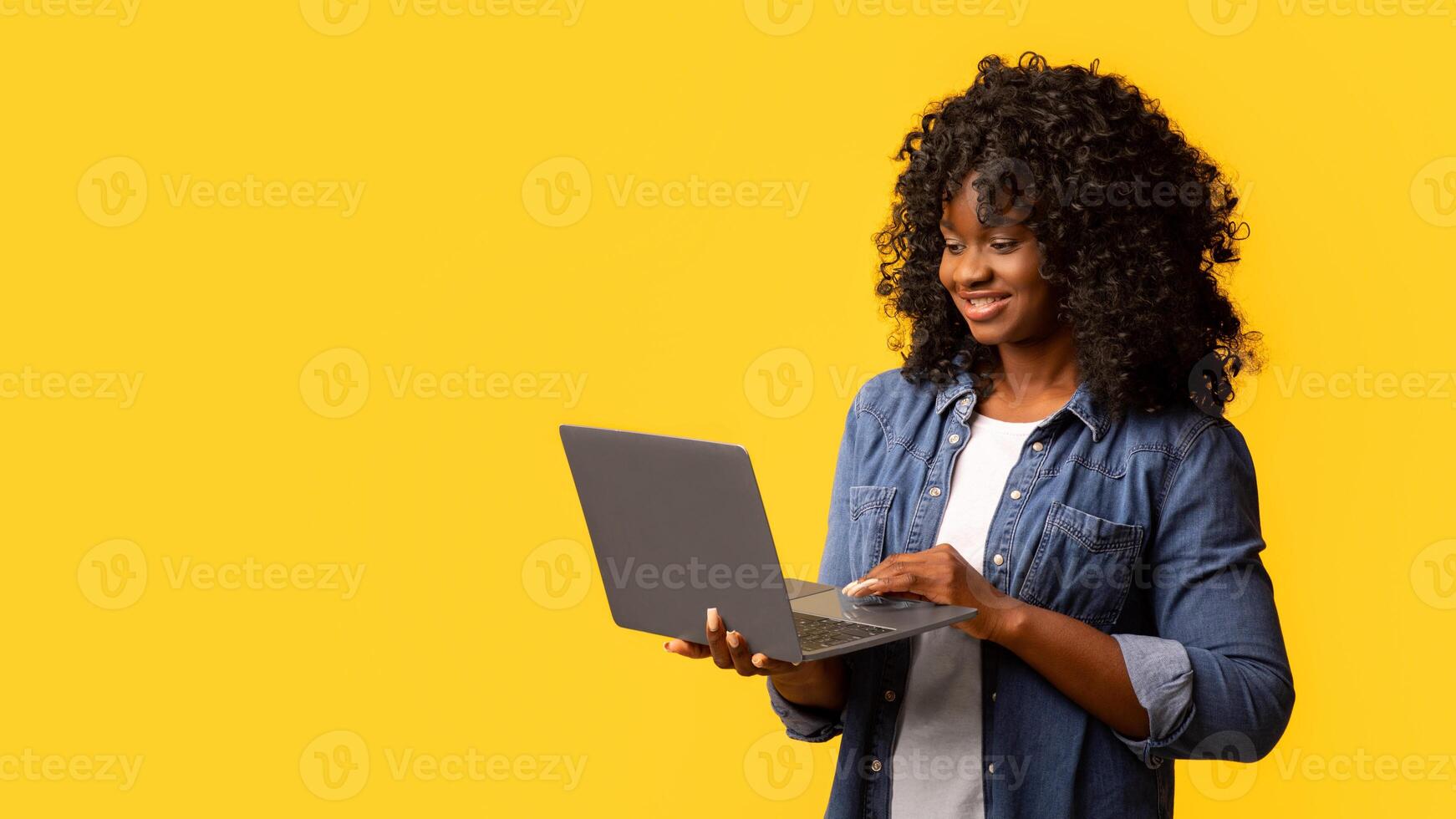 Pretty young black lady using modern laptop on yellow studio background, copy space. Cute african american curly woman chatting with friends or checking webstores. Modern technologies concept photo