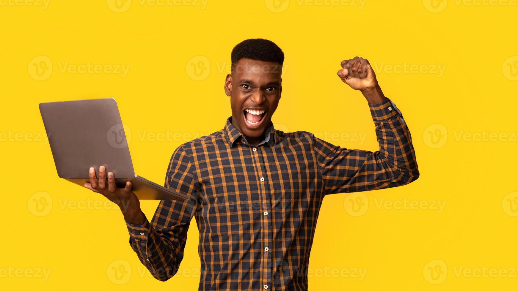 Euphoric Black Gguy Celebrating Success With Laptop, Raising Clenched Fist And Exclaiming With Excitement, Joyful African American Man Holding Computer, Standing On Yellow Background, Copy Space photo