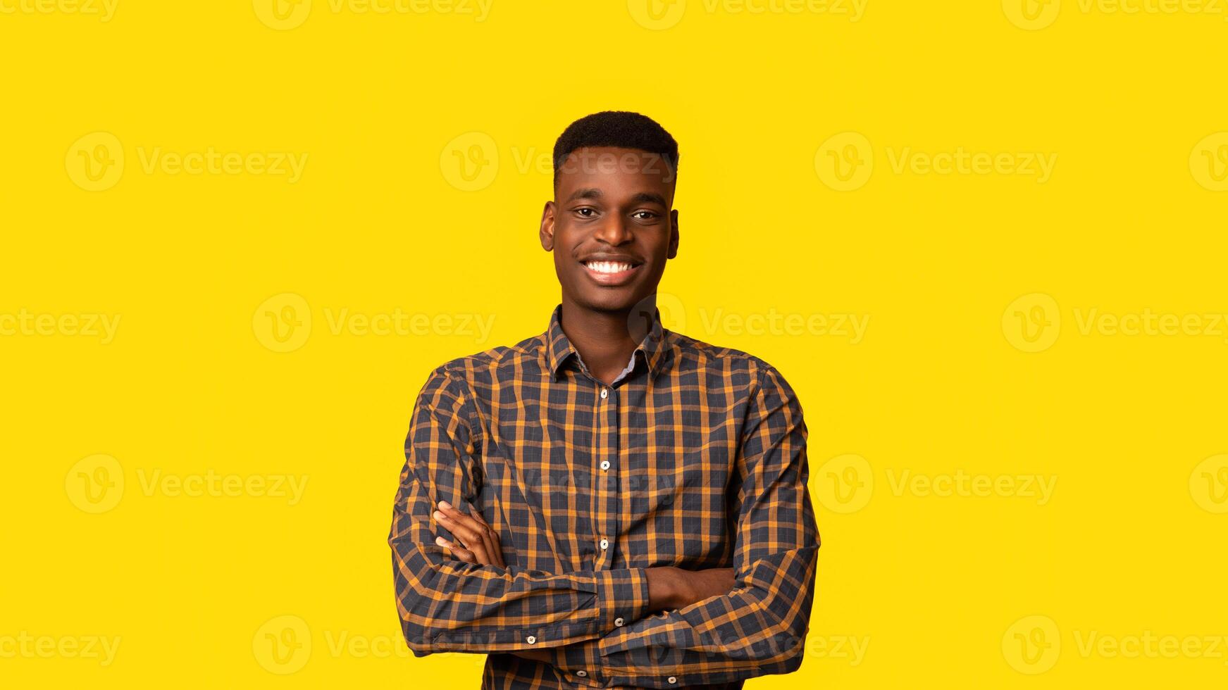 Positive Young Black Guy Posing With Folded Arms Over Yellow Studio Backgound, Handsome Millennial African American Man Wearing Checkered Shirt, Smiling And Looking At Camera, Panorama With Copy Space photo