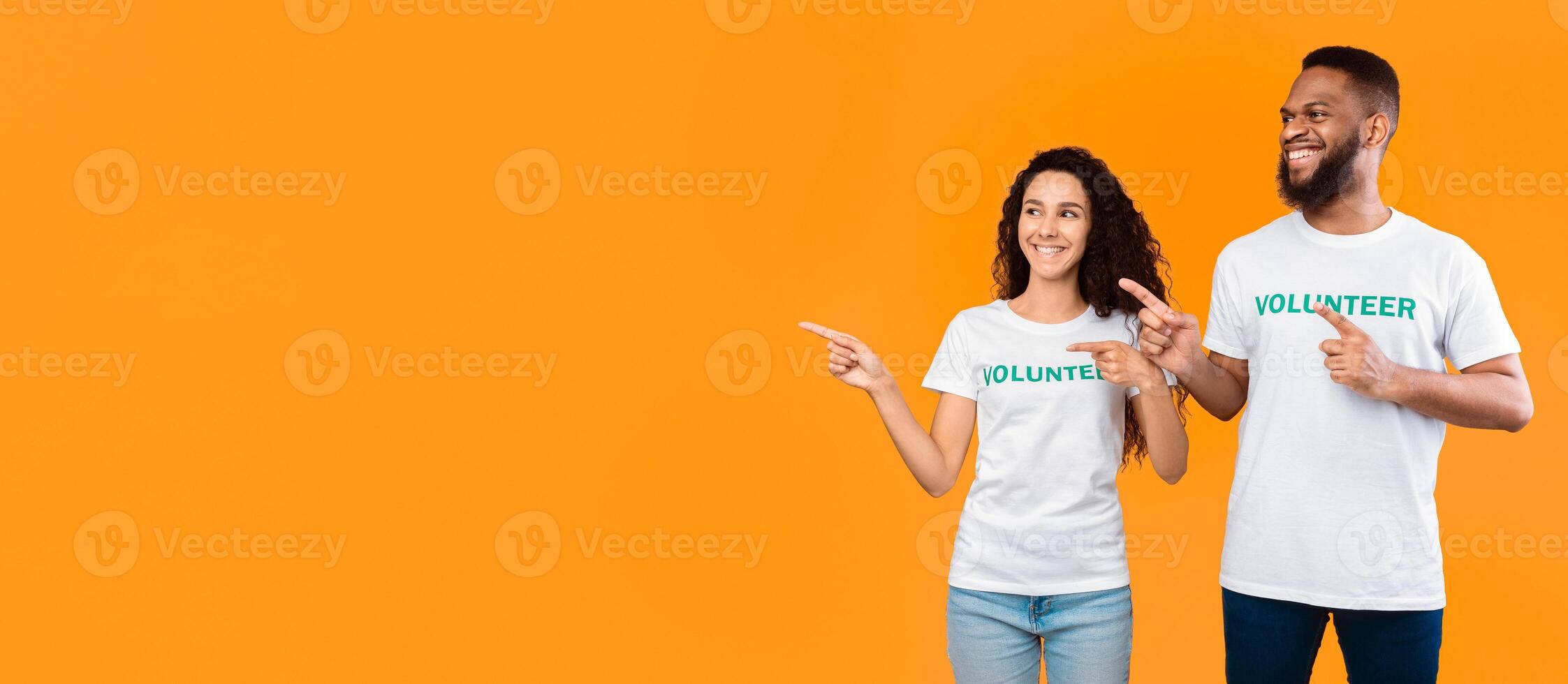 Look Aside. Two Multiethnic Volunteers Pointing Fingers At Free Space For Your Text Posing Standing Over Yellow Studio Background. Volunteering And Charity Advert. Panorama photo