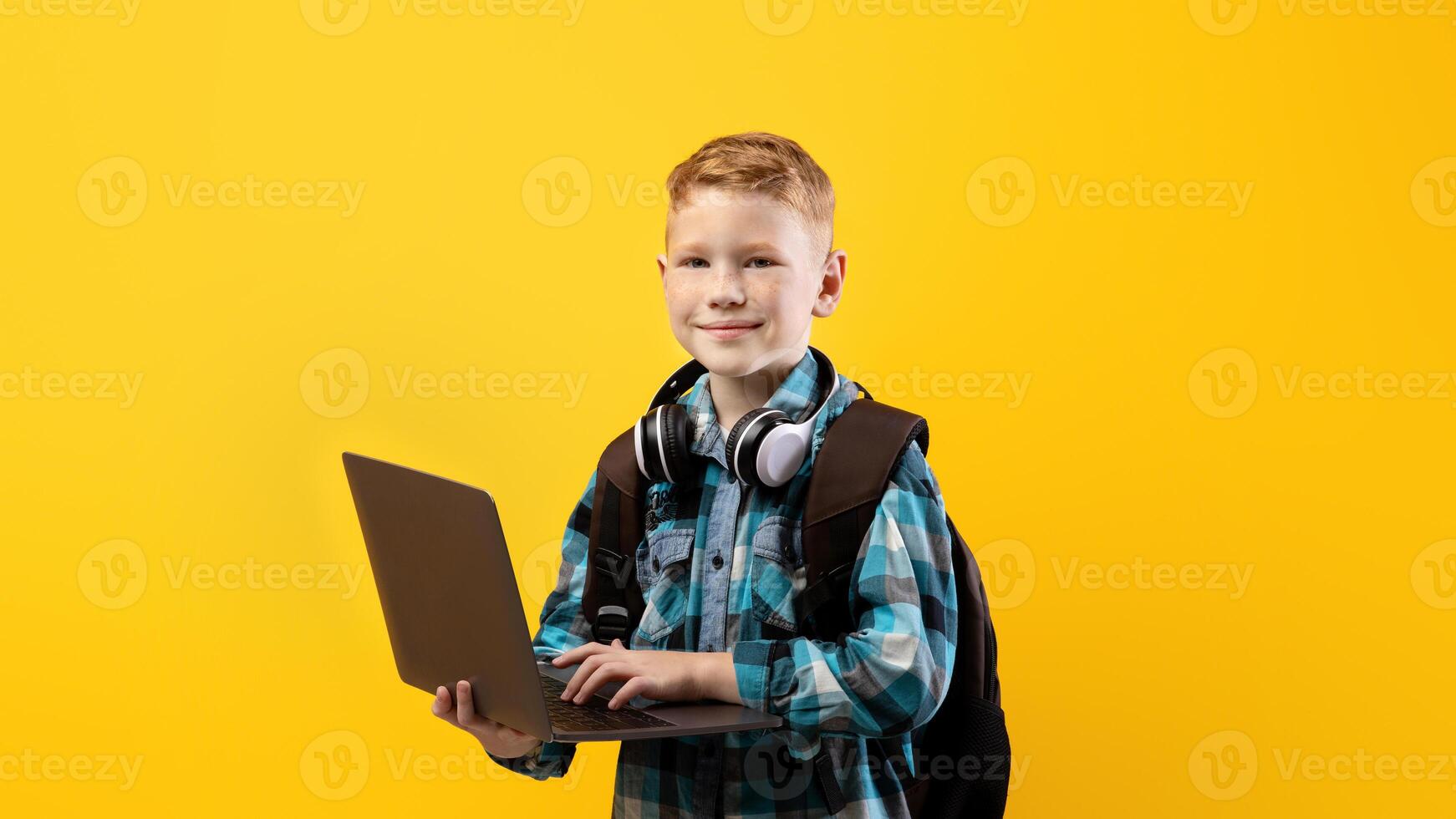 Technologies in school education. Cheerful schoolboy with backpack and headset holding laptop over yellow studio background, copy space. Cute kid using laptop for studying, doing homework photo