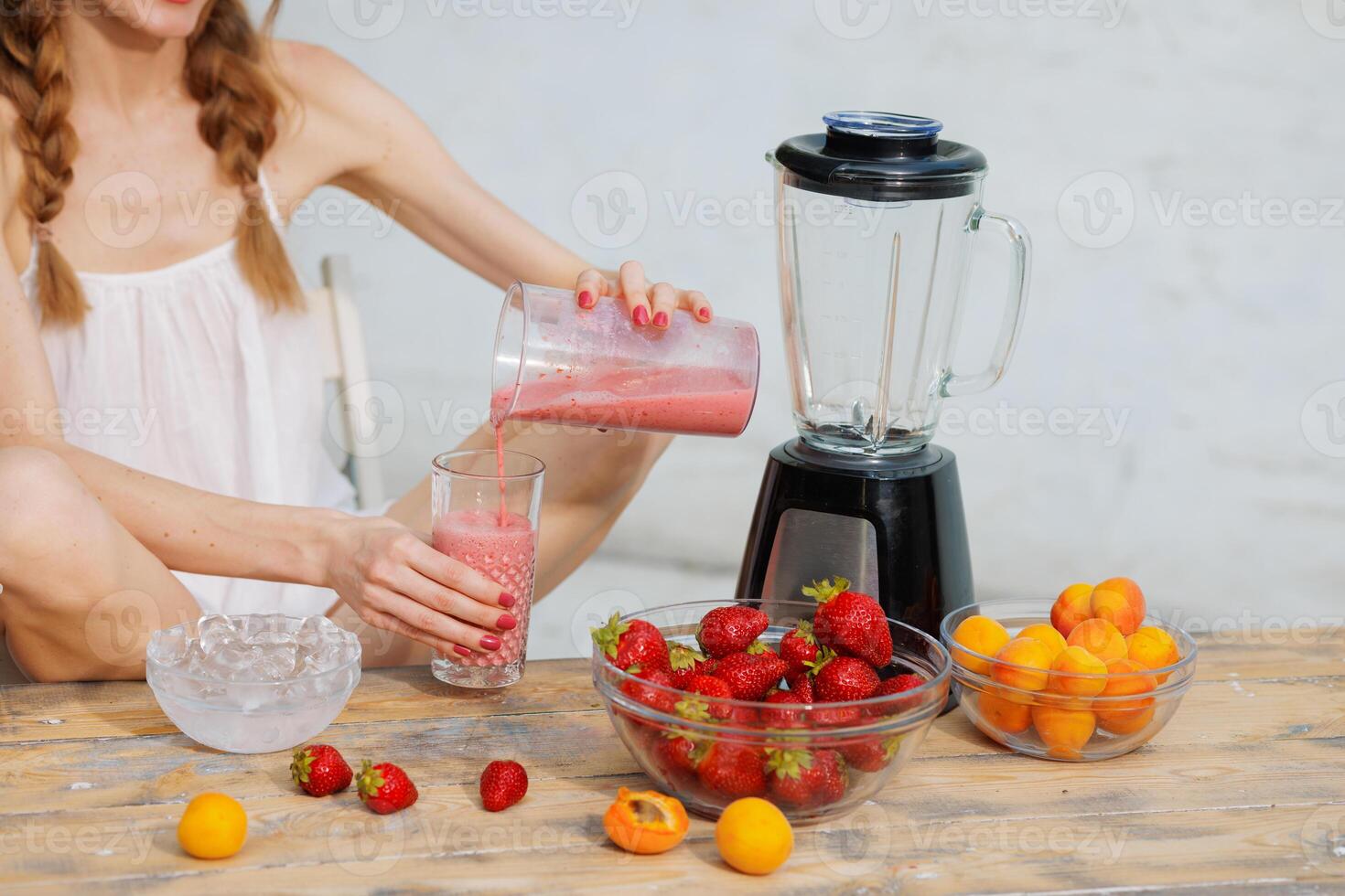 Woman pouring fresh homemade strawberry smoothie into a glass from blender cup on rustic wooden table with bowls of strawberries, apricots, and ice. Healthy summer drink concept. photo