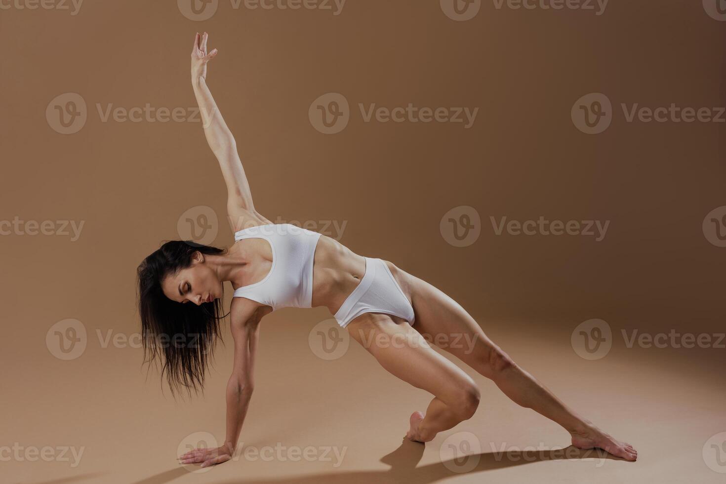 Fit woman in white activewear performing a dynamic yoga pose on a brown studio background, demonstrating strength, balance, and body control in minimalist wellness concept. photo