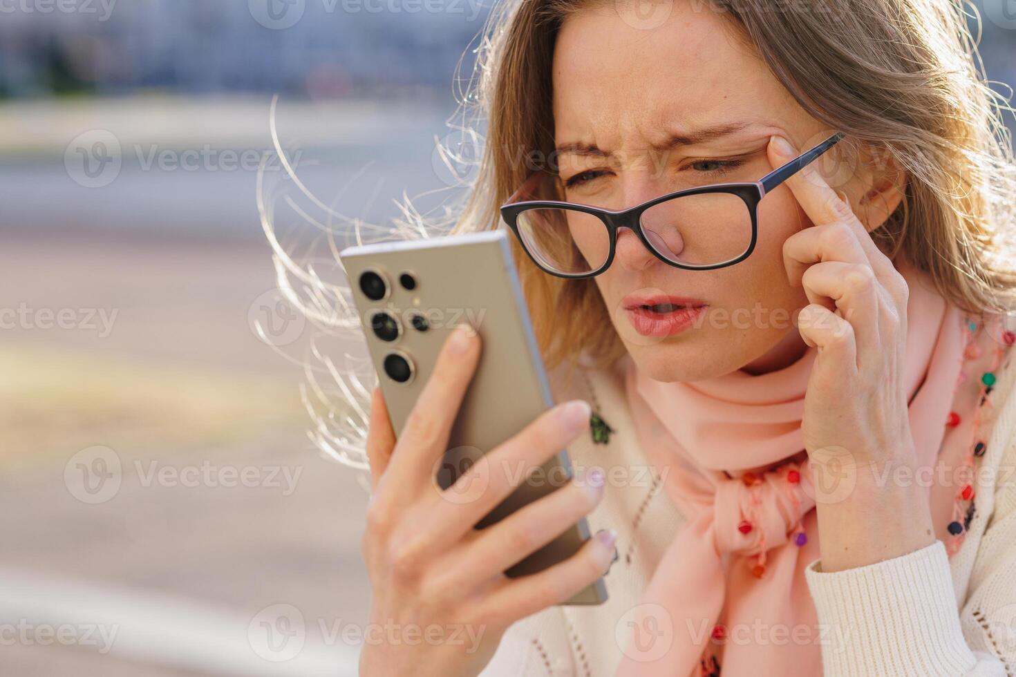 Frustrated woman squinting at smartphone screen while wearing glasses, symbolizing vision problems, digital eye strain, and poor eyesight in bright outdoor conditions. photo
