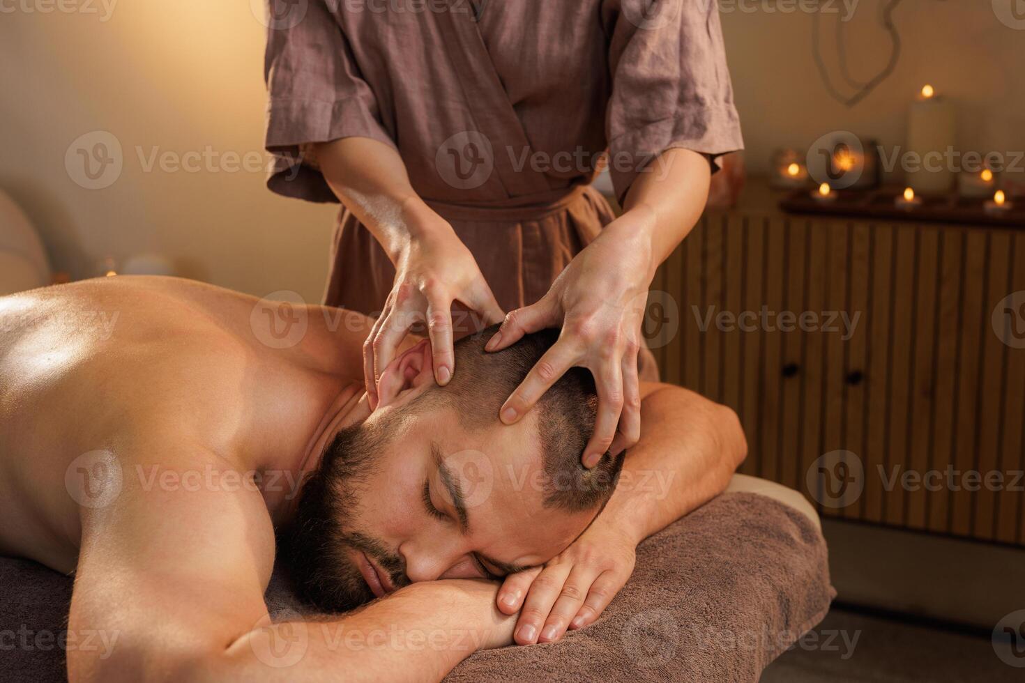 Professional therapist performing a relaxing head massage on a bearded man lying on a spa table in a calm, candle-lit wellness room with warm and peaceful atmosphere. photo