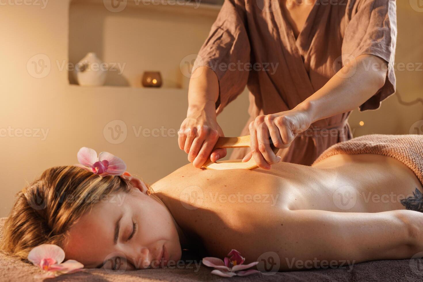 Spa therapist performs wooden tool back massage to relieve muscle tension and promote relaxation in a tranquil environment decorated with orchids and warm ambient lighting. photo