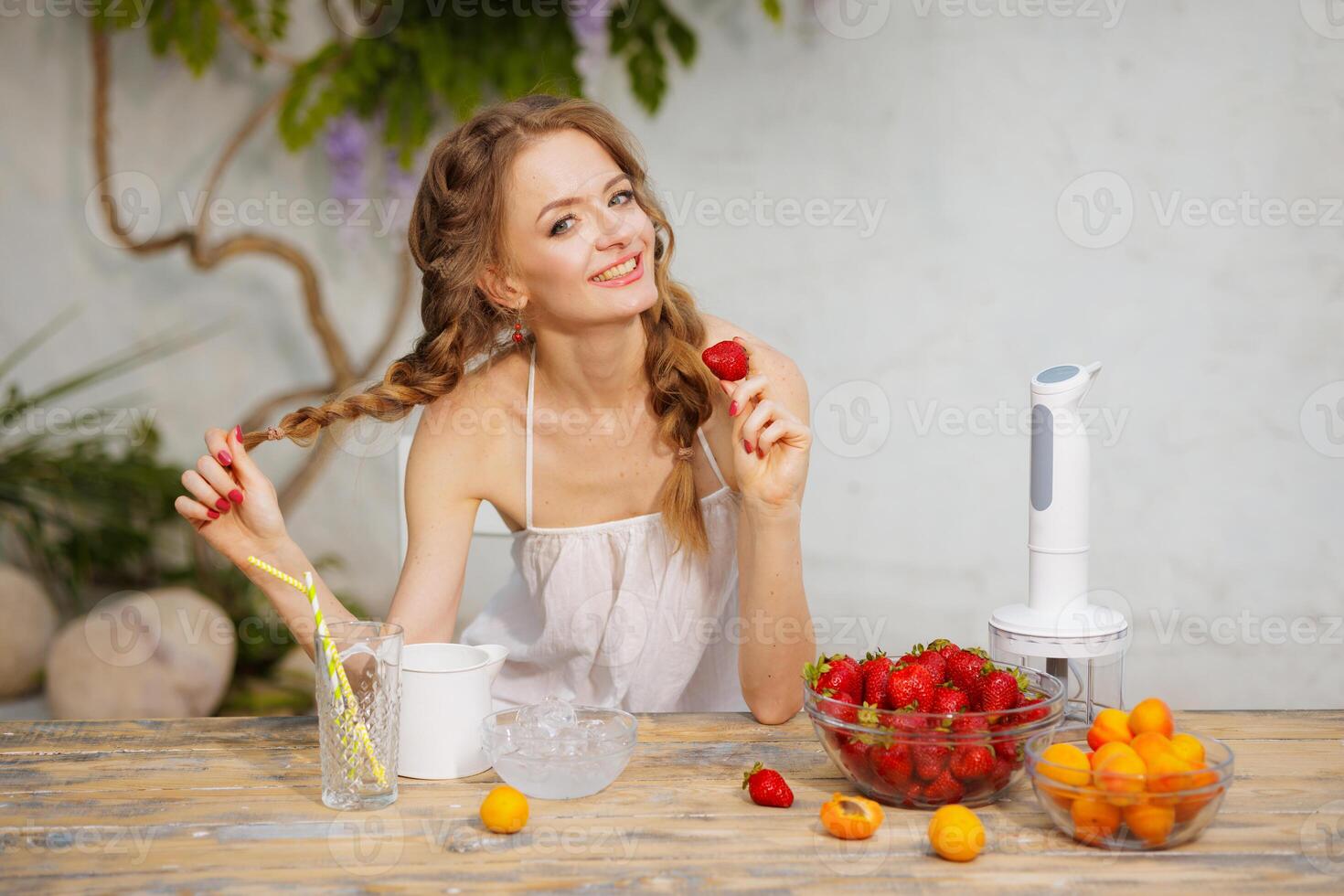 Smiling young woman with braided hair holding a strawberry at a rustic wooden table with fresh fruits, ice, and a blender in a summer garden, evoking health and joy. photo