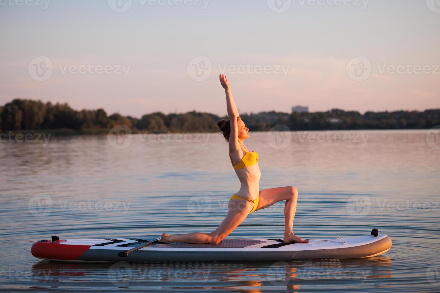 Woman in yellow bikini practicing yoga on paddleboard in warrior pose at sunrise, balancing on calm water, expressing focus, strength, and harmony with nature. photo