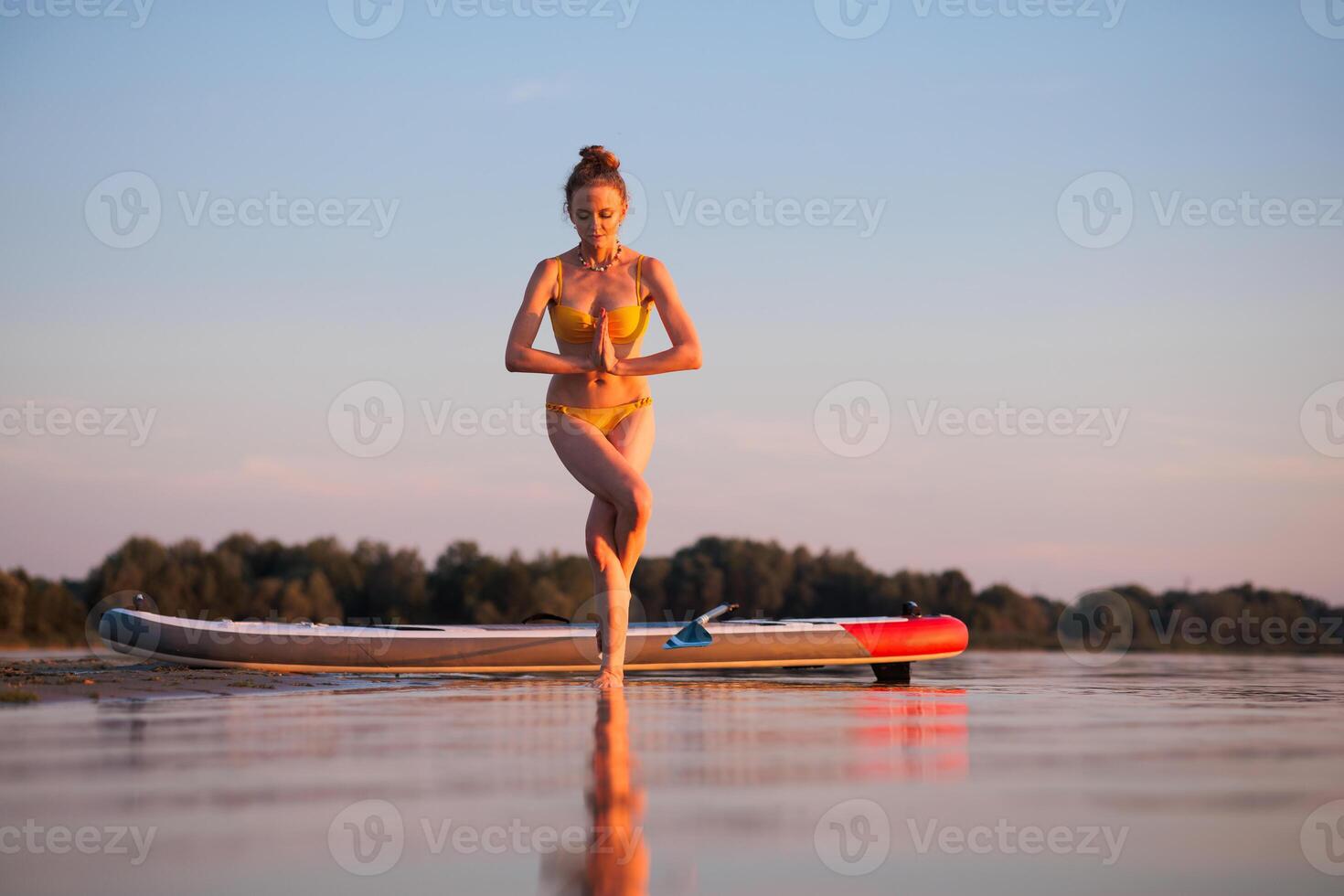Woman in yellow bikini practicing yoga in tree pose on shallow water beside paddleboard at sunrise, expressing balance, mindfulness, and connection to nature. photo