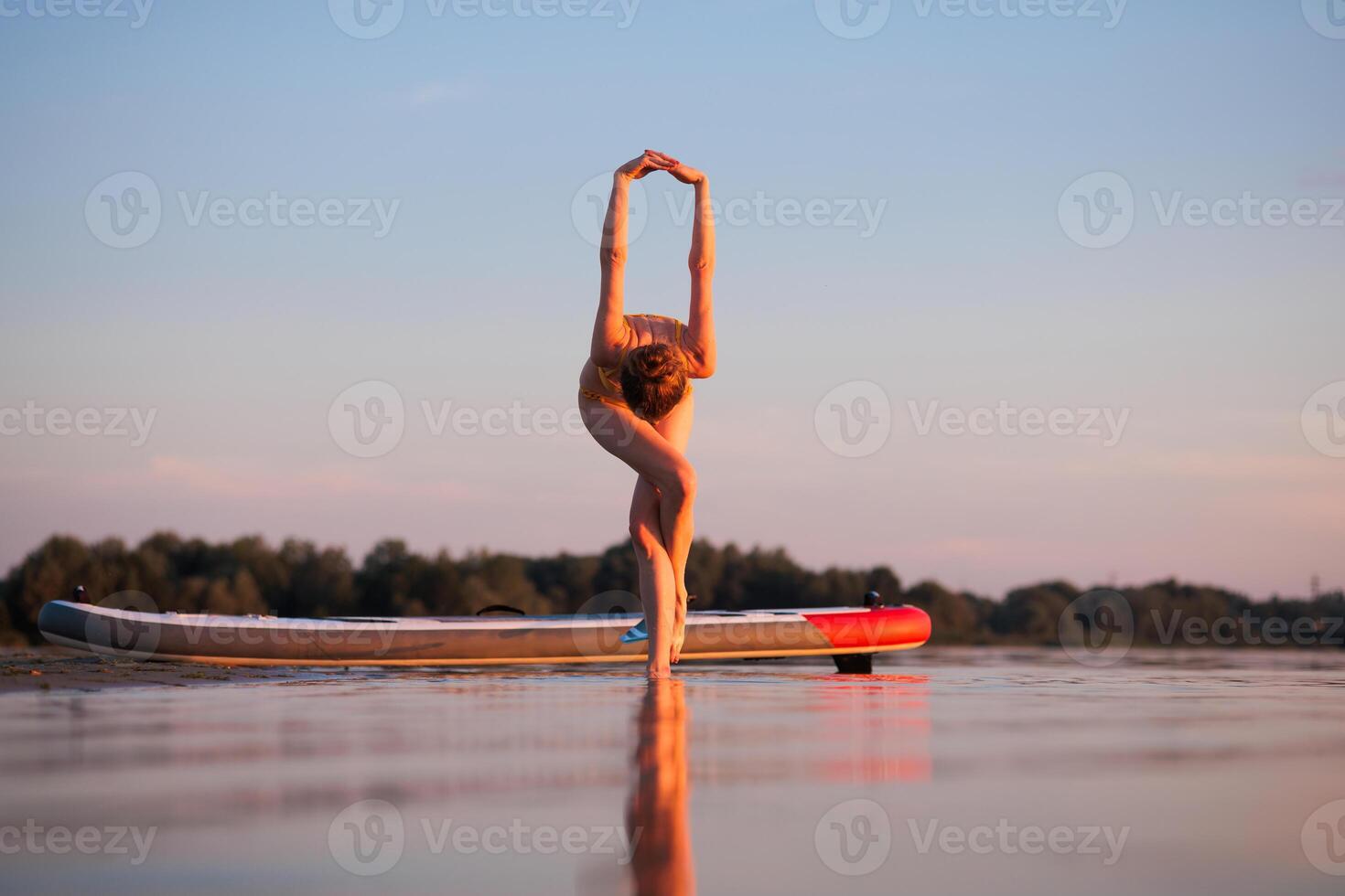 Woman in yellow bikini balancing in standing forward yoga pose on one leg beside paddleboard at sunset, expressing flexibility, focus, and body control over calm water. photo