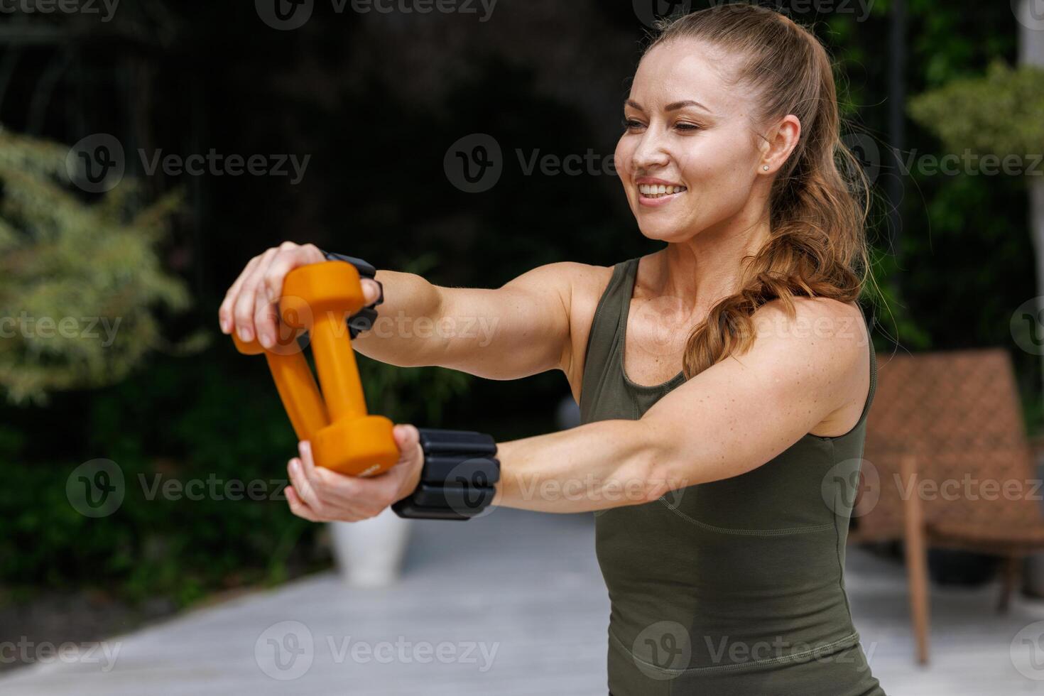 Smiling athletic woman in green sportswear holding orange dumbbells during an outdoor workout, showing strength, positivity, and motivation for a healthy and active lifestyle. photo