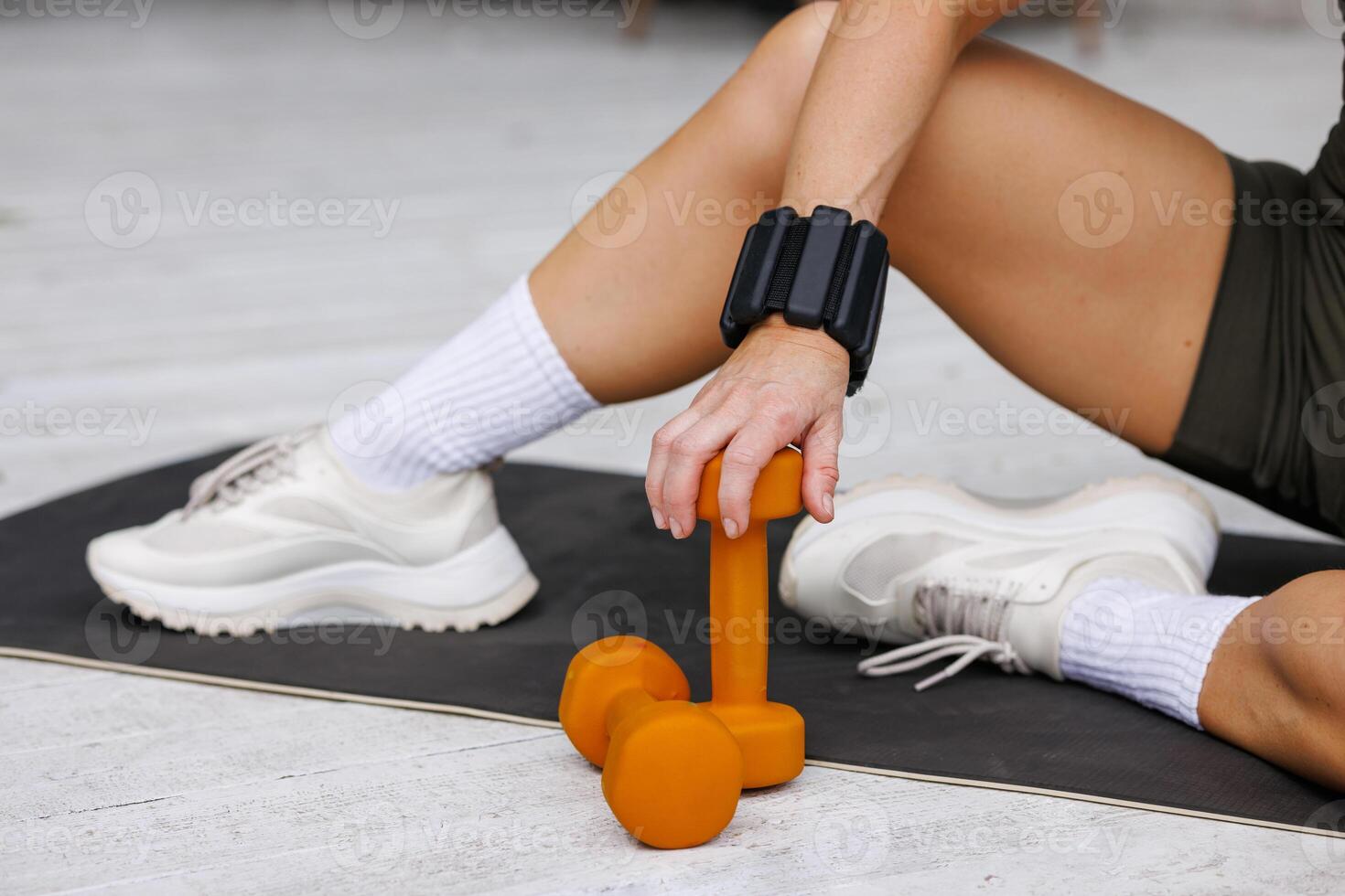 Close up of a womans hand grasping an orange dumbbell on a yoga mat outdoors, with white sneakers and wrist weights, highlighting active fitness, strength training, and a healthy lifestyle. photo