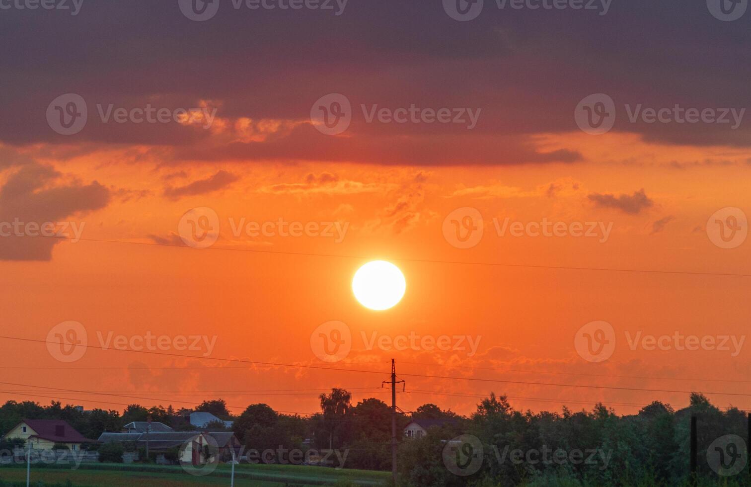 A stunning sunset paints the sky in shades of orange and purple over a tranquil countryside. Silhouettes of trees and houses dot the landscape, creating a serene atmosphere photo