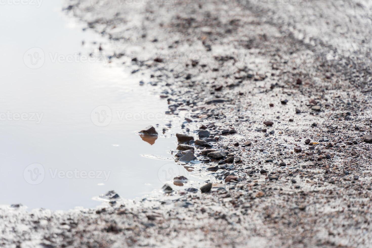 A rural gravel road shows a mixture of wet stones and puddles after recent rainfall, capturing reflections of a cloudy sky during early morning light photo