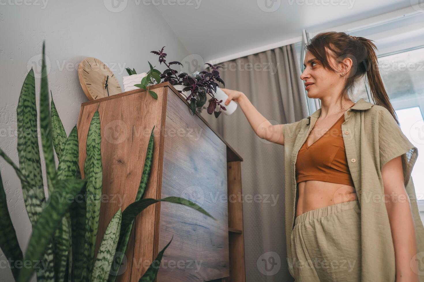 woman with tied brown hair spray houseplants from behind in softly lit room with wooden cabinet, potted flowers, home wellness, indoor gardening, sustainable lifestyle photo