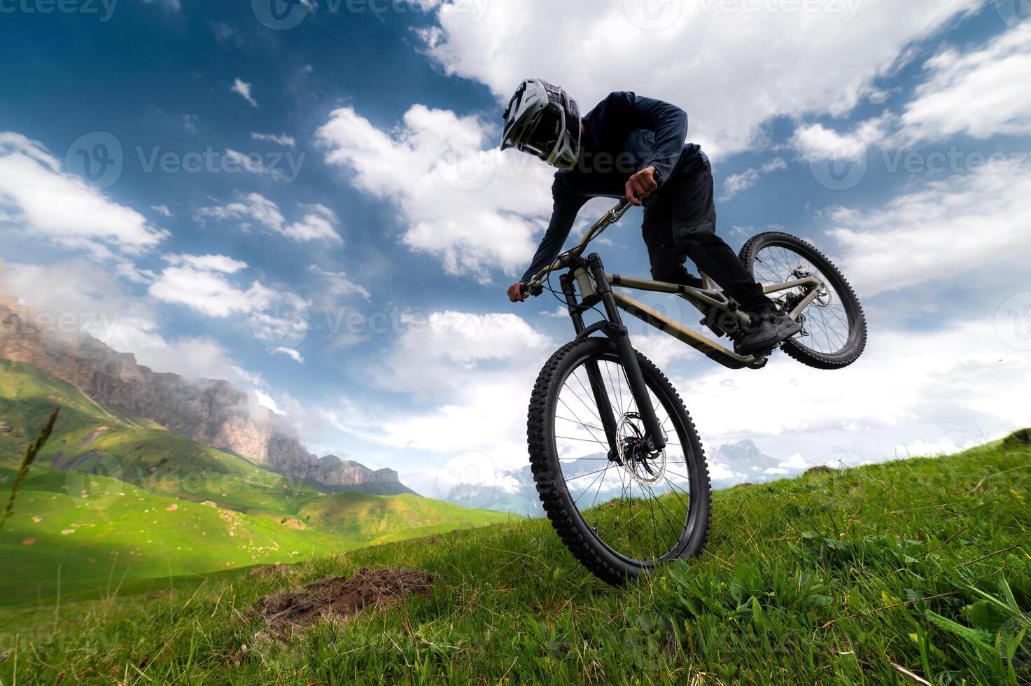 mountain biker takes off with his rear wheel leaning on his front wheel, performing a manuel or wheelie trick, against a mountain landscape in the fields in summer photo