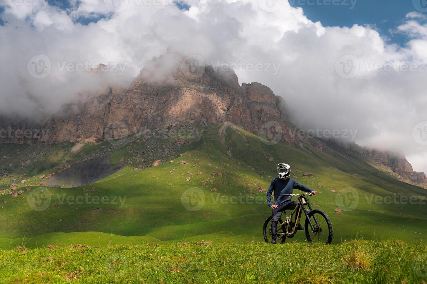 View of cyclist standing with mountain bike in summer green fields in Dolomites with high rocky mountain in background in thick clouds on sunny day. Off-piste riding photo