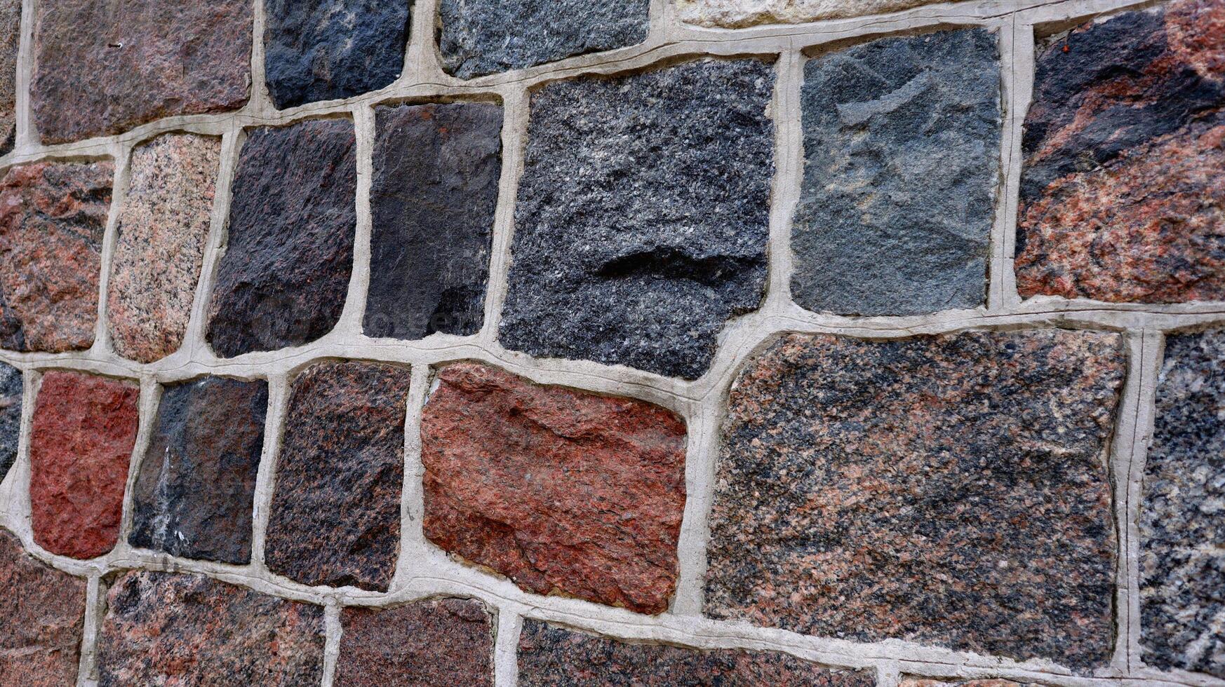 Close-up view of a multicolored stone wall with irregular rectangular blocks and white grout. The textured surface and natural colors give a rustic and architectural feel. photo