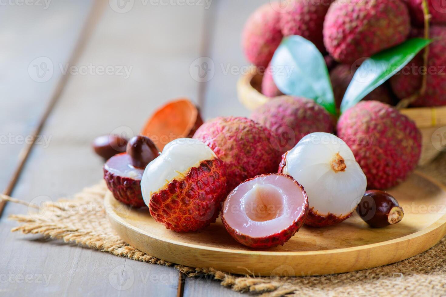 fresh ripe lychees in a bamboo basket with green leaves on a rustic wooden table. One peeled lychee reveals the juicy white flesh inside.. photo