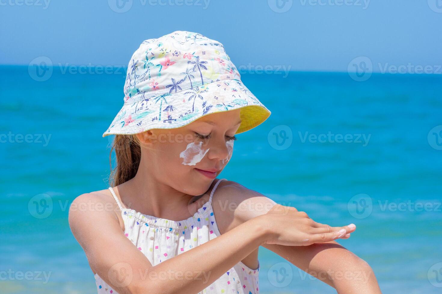 Child is applying sunscreen on her arm at the beach, wearing a colorful hat, with bright ocean waves in the background, emphasizing sun protection and enjoyment photo