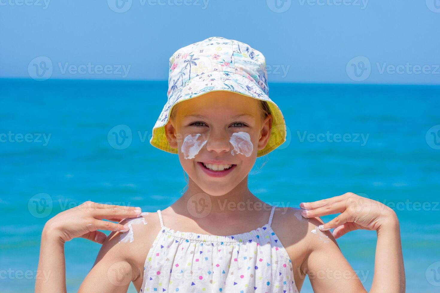 Child happily applying sunscreen on her face and shoulders at the beach, wearing a vibrant hat, with the ocean waves and bright sky creating a joyful summer atmosphere photo