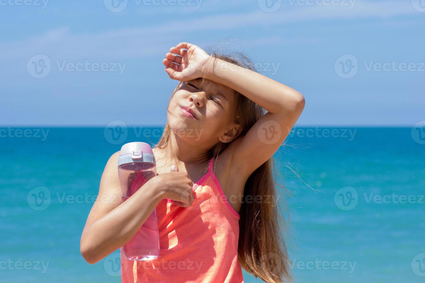 Child with closed eyes holding bottle of water and her arm on forehead because of hotness in summer over blue sea photo